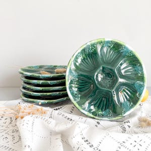 Peut inclure: Ensemble de six assiettes à huîtres en céramique verte avec une finition brillante. Les assiettes présentent un design festonné avec des compartiments individuels. Une assiette est dressée, montrant l'intérieur détaillé. Les assiettes sont sur une nappe en dentelle blanche.