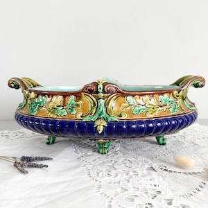 May include: A ceramic decorative bowl with a blue, brown, and green colour scheme. The bowl has a detailed, ornate design with handles and is supported by small feet. The rim is blue, and the body features a textured pattern.