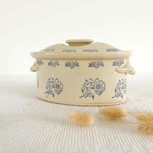 May include: Cream-coloured ceramic casserole dish with a lid, decorated with blue floral patterns. The dish features two handles and a knob on the lid. The floral design is repeated around the dish's body.