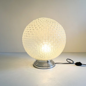 Peut inclure: Une lampe de table en verre blanc avec un motif géométrique et une base chromée. La lampe est allumée et la lumière brille à travers le verre.