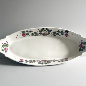 May include: An oval, white ceramic serving platter with a decorative border of green leaves, red berries, and pink flowers. The platter has scalloped edges and is suitable for serving food.