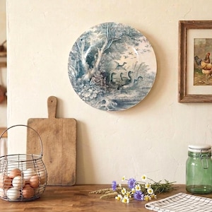 Puede incluir: Un plato de pared redondo de cerámica con un diseño azul y blanco que muestra patos nadando en un estanque cerca de árboles. El plato se exhibe en una pared color crema junto a un cuadro enmarcado de pollos y varios utensilios.