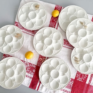 Puede incluir: Ocho platos de cerámica blanca para ostras, cada uno con seis compartimentos en forma de concha y un pozo central. Están colocados sobre un mantel a rayas blancas y rojas, con dos limones amarillos. Un plato de servicio clásico.