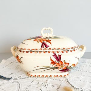 May include: Cream-coloured, oval serving dish with a lid, featuring a floral design in shades of orange, red, and green. The dish has a decorative handle on the lid and side handles. The base is rectangular. A vintage kitchenware item.