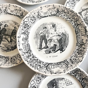 K&ouml;nnte beinhalten: Ein Set antiker Keramikteller mit Schwarzwei&szlig;illustrationen. Die Teller zeigen Szenen von Menschen, die W&auml;sche waschen, in einer Fabrik arbeiten und andere allt&auml;gliche Aktivit&auml;ten ausf&uuml;hren. Die Teller haben einen dekorativen Rand mit floralen Mustern.