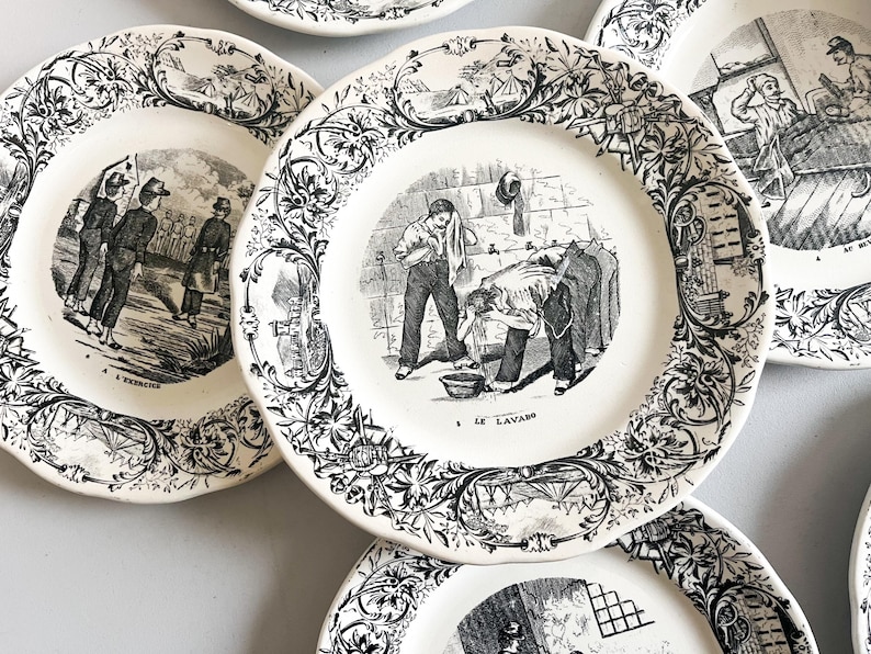 K&ouml;nnte beinhalten: Ein Set antiker Keramikteller mit Schwarzwei&szlig;illustrationen. Die Teller zeigen Szenen von Menschen, die W&auml;sche waschen, in einer Fabrik arbeiten und andere allt&auml;gliche Aktivit&auml;ten ausf&uuml;hren. Die Teller haben einen dekorativen Rand mit floralen Mustern.
