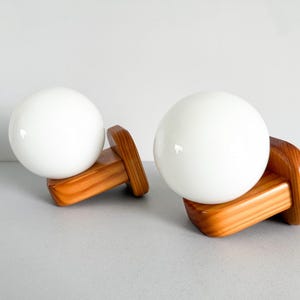 May include: A pair of wall-mounted light fixtures featuring spherical white glass globes attached to rectangular, light-brown wooden bases with visible wood grain patterns. The lamps are designed with a smooth, rounded aesthetic.