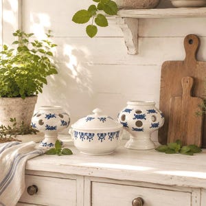 Könnte beinhalten: Ein Set aus weißem Keramikgeschirr mit blauem Blumenmuster, bestehend aus zwei bauchigen Vasen mit kreisförmigen Aussparungen und einer Terrine mit Deckel, arrangiert auf einer weißen Oberfläche neben einem gestreiften Tuch.