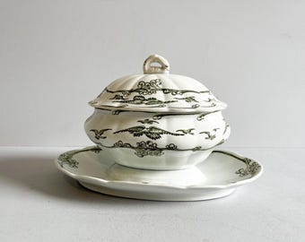 May include: A white porcelain gravy boat with a lid, featuring a green and white floral design. The boat has a pedestal base and a matching saucer.