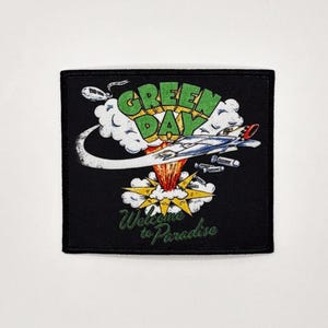 Può includere: Toppa quadrata nera con una grafica di un jet, un'esplosione e le parole "GREEN DAY" in verde e giallo. Il testo "Welcome to Paradise" è incluso in verde. La toppa ha un bordo nero.