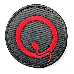 Puede incluir: Parche redondo negro con un diseño bordado en rojo. El diseño presenta una letra "Q" roja estilizada con una cola de serpiente. El parche tiene un borde negro, probablemente un emblema o logotipo.