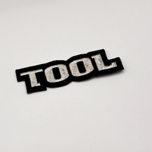 K&ouml;nnte beinhalten: Schwarzer, gestickter Aufn&auml;her mit dem Wort "TOOL" in silbernem Faden. Der Aufn&auml;her hat einen schwarzen Rand und eine rechteckige Form. Der Text ist fett und ohne Serifen.