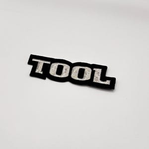 K&ouml;nnte beinhalten: Schwarzer, gestickter Aufn&auml;her mit dem Wort "TOOL" in silbernem Faden. Der Aufn&auml;her hat einen schwarzen Rand und eine rechteckige Form. Die Buchstaben sind in einer fetten, serifenlosen Schriftart.
