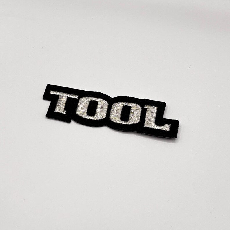 K&ouml;nnte beinhalten: Schwarzer, gestickter Aufn&auml;her mit dem Wort "TOOL" in silbernem Faden. Der Aufn&auml;her hat einen schwarzen Rand und eine rechteckige Form. Die Buchstaben sind in einer fetten, serifenlosen Schriftart.