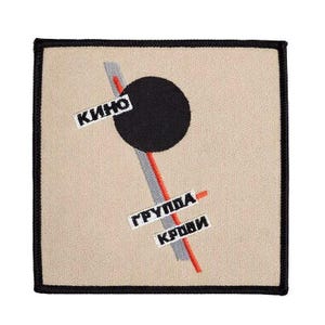 May include: A square embroidered patch with a beige background and a black border. The design features a large black circle, a diagonal gray and red stripe, and white rectangular labels with black Cyrillic text reading KINO and GRUPPA KROVI.