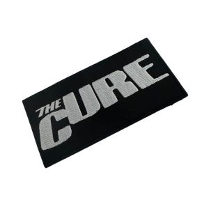 Könnte beinhalten: Ein rechteckiger, schwarz bestickter Aufnäher mit dem Text THE CURE in fetten, weißen Großbuchstaben. Die Stickerei weist ein sichtbares, strukturiertes Stichmuster auf den Buchstaben auf, das sich vom schwarzen Stoffhintergrund abhebt.