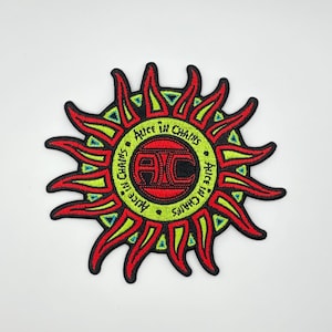 Op de afbeelding: Geborduurde patch met het Alice in Chains logo. Het ontwerp is een gestileerde zonnestraal in rood, groen en zwart. De naam van de band staat rond het centrale logo, dat de initialen "AC" bevat.