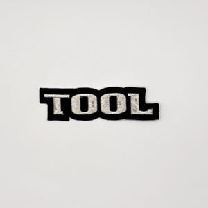 K&ouml;nnte beinhalten: Schwarzer, gestickter Aufn&auml;her mit dem Wort "TOOL" in silbernem Faden. Der Aufn&auml;her hat einen schwarzen Rand und befindet sich auf einem wei&szlig;en Hintergrund. Die Buchstaben sind im Blockstil.