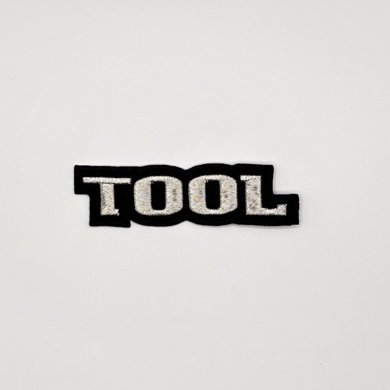 K&ouml;nnte beinhalten: Schwarzer, gestickter Aufn&auml;her mit dem Wort "TOOL" in silbernem Faden. Der Aufn&auml;her hat einen schwarzen Rand und befindet sich auf einem wei&szlig;en Hintergrund. Die Buchstaben sind im Blockstil.