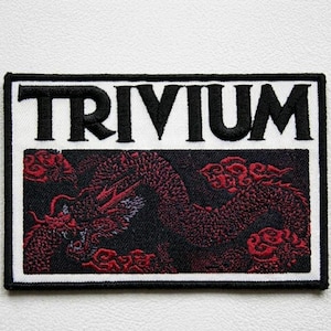Könnte beinhalten: Gestickter Aufnäher mit dem Wort "TRIVIUM" in Schwarz über einem roten Drachendesign auf schwarzem Hintergrund. Der Aufnäher hat einen weißen Rand und ist rechteckig.