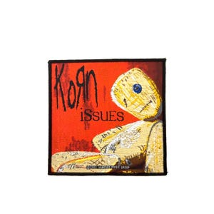 Op de afbeelding: Een vierkante stoffen patch met een rode achtergrond en de tekst KoRn en iSSUES in het zwart. Een gele lappenpop met één blauw knoopoog staat aan de rechterkant afgebeeld. De onderrand bevat de tekst 2022 WARNER MUSIC GROUP.