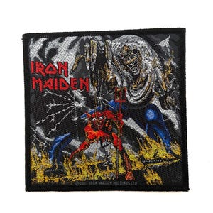 Puede incluir: Un parche bordado cuadrado con el logotipo de Iron Maiden en texto rojo. El gráfico muestra una figura esquelética grande y pálida sobre un personaje rojo más pequeño parecido a un diablo que sostiene un tridente entre llamas amarillas.
