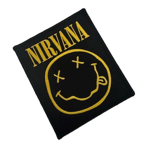 May include: A rectangular black fabric patch featuring yellow embroidered text that reads NIRVANA above a yellow embroidered smiley face graphic with X-shaped eyes and a wavy mouth, all stitched onto a solid black background material.