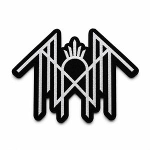 May include: A black and white embroidered patch featuring a symmetrical geometric design. The emblem consists of sharp, angular lines forming a house-like structure with a central arched element topped by a small, radiating sunburst motif.