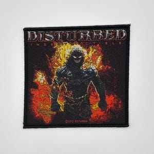 May include: Black square patch with the word "DISTURBED" in white, above "INDESTUCTIBLE". A demonic figure is in the centre, surrounded by flames. The patch has a black border and the text "©2012 DISTURBED" at the bottom.