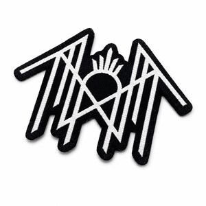 May include: A black and white embroidered patch featuring a stylized, angular geometric design. The graphic consists of sharp, interconnected lines forming a symmetrical shape with a central sunburst element, all outlined in a thick black border.