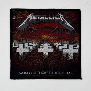 Puede incluir: Un parche de tela tejido cuadrado que presenta el logotipo de Metallica en blanco en la parte superior, sobre una escena de color rojo oscuro y negro de cruces blancas en un campo. El texto MASTER OF PUPPETS está impreso en blanco en la parte inferior.