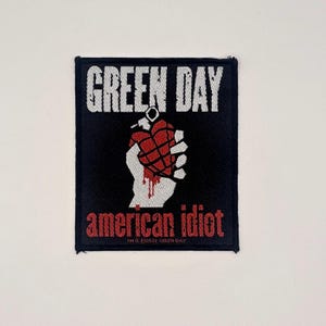Può includere: Toppa quadrata nera con la scritta bianca "GREEN DAY" in alto. Sotto, una grafica di una mano bianca che tiene una granata rossa a forma di cuore. Il testo "american idiot" è in rosso in basso.