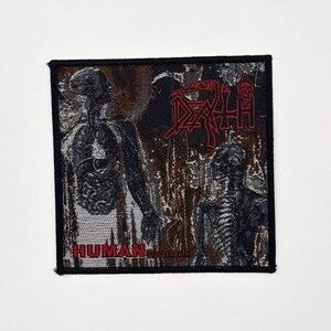 Op de afbeelding: Vierkante patch met een zwarte rand met een skeletfiguur en het woord "DEATH" in het rood. De tekst "HUMAN..." is onderaan zichtbaar. De achtergrond heeft een donkere, getextureerde uitstraling.