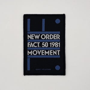 Puede incluir: Un parche rectangular negro con el texto "NEW ORDER FACT. 50 1981 MOVEMENT" en blanco y azul. El parche tiene líneas verticales azules y un círculo azul. El texto "©2023 VITALTURN" está en la parte inferior.