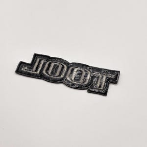 K&ouml;nnte beinhalten: Ein schwarz-silberner, gestickter Aufn&auml;her mit dem Wort "JOOT" in einer stilisierten Schriftart. Der Aufn&auml;her hat eine rechteckige Form mit abgerundeten Kanten und ist zum Aufn&auml;hen oder Aufb&uuml;geln auf Stoff gedacht.