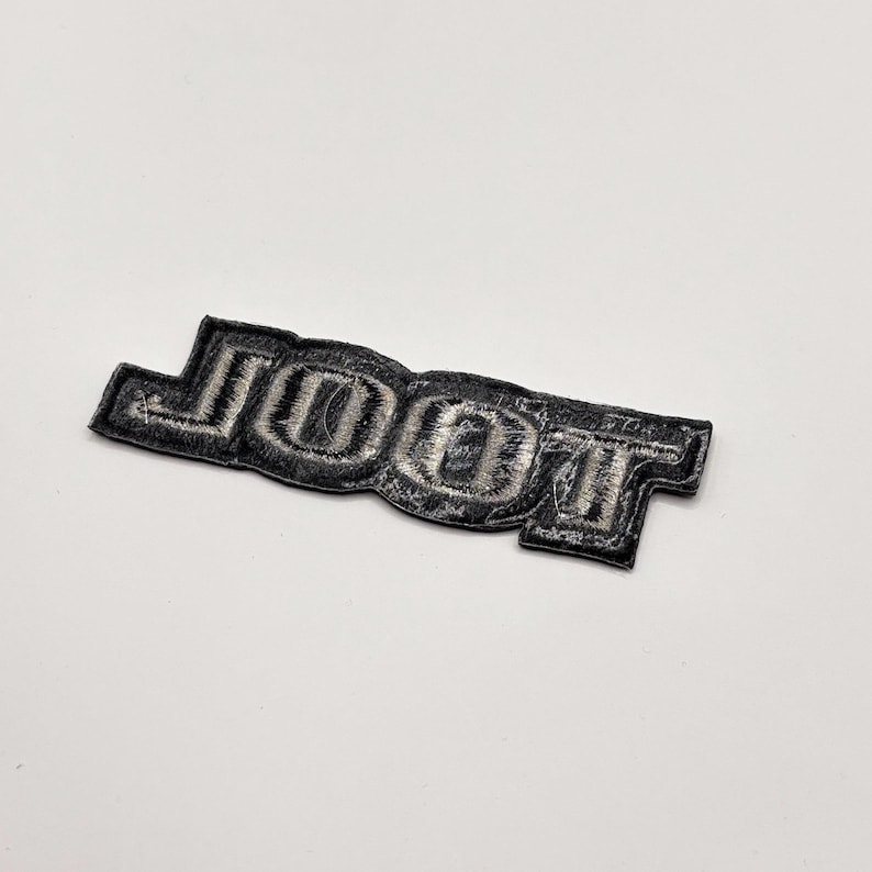 K&ouml;nnte beinhalten: Ein schwarz-silberner, gestickter Aufn&auml;her mit dem Wort "JOOT" in einer stilisierten Schriftart. Der Aufn&auml;her hat eine rechteckige Form mit abgerundeten Kanten und ist zum Aufn&auml;hen oder Aufb&uuml;geln auf Stoff gedacht.