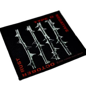 May include: A square black fabric patch featuring four vertical silver barbed wire designs. The text TYPE O NEGATIVE is printed in red at the top, with RUST and OCTOBER in red at the bottom, and copyright text in the bottom right corner.