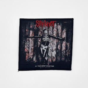 Puede incluir: Parche cuadrado negro con una figura de esqueleto con los brazos extendidos sobre un fondo de líneas verticales, similares a madera desgastada. La palabra "Slipknot" está en rojo en la parte superior. Debajo de la figura, el texto dice "5: THE GRAY CHAPTER" y "©2013 SLIPKNOT."