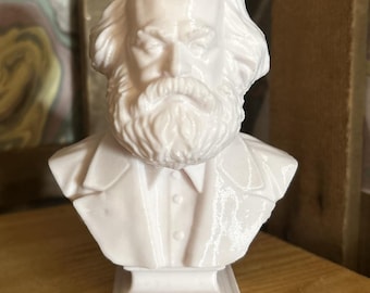 May include: A white bust sculpture of a man with a full beard and long hair. The sculpture is of a man wearing a collared shirt and jacket. The bust is mounted on a rectangular base.