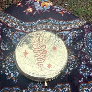 Op de afbeelding: Een goudkleurige compacte spiegel met een rood slang- en sterontwerp. De ronde spiegel reflecteert de omgeving. De spiegel ligt op een zwart-blauw patroon textiel.