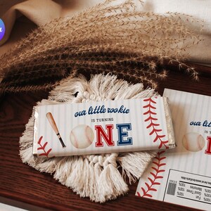 May include: A baseball-themed candy bar wrapper with the text "our little rookie is turning ONE" and a baseball bat and ball graphic. The wrapper is on a macrame coaster and a wooden tray.