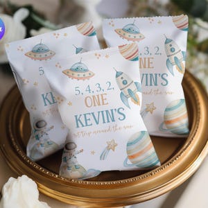 Puede incluir: Tres bolsas blancas para golosinas con temática espacial, con cohetes, planetas y ovnis. Las bolsas tienen el texto "5, 4, 3, 2, ONE KEVIN'S 1st trip around the sun" impreso. Las bolsas están sobre una bandeja dorada.