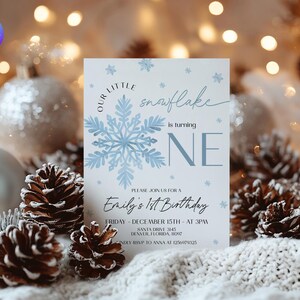 May include: A light blue and white birthday invitation with snowflake graphics. The invitation reads "Our Little Snowflake is turning ONE" and includes event details. The invitation is surrounded by pine cones and ornaments.