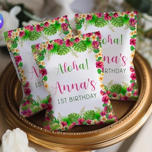 Könnte beinhalten: Drei weiße Partygeschenktüten mit tropischem Blumendesign, darunter Hibiskusblüten und Monstera-Blätter. Auf den Tüten steht "Aloha! Anna's 1st Birthday" in grüner und rosa Schrift. Sie werden auf einem goldfarbenen Tablett präsentiert.