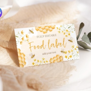 Può includere: Un'etichetta alimentare piegata con un design ad acquerello con favi, api e fiori gialli. L'etichetta recita "Food label" in un carattere corsivo, con spazio per aggiungere testo. Lo sfondo è di un colore morbido e neutro.