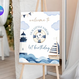 Puede incluir: Un letrero de cumpleaños con temática náutica sobre un caballete. El letrero presenta un faro, un velero y un salvavidas con una gaviota. El texto dice "Bienvenido a Nuestro Pequeño Marinero 1er Cumpleaños" en azul y dorado.
