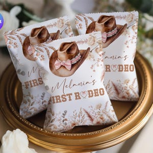 May include: Three white party favor bags with a brown cowboy hat illustration and the text "Melania's FIRST RODEO". The bags are decorated with floral elements and a pink bow. The bags are on a gold-colored tray.