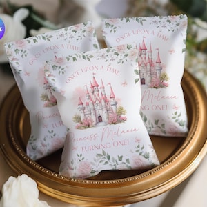 May include: Three white party favor bags with a pink castle design, floral accents, and the text "ONCE UPON A TIME" and "Princess Melania TURNING ONE!". The bags are arranged on a gold-colored tray.