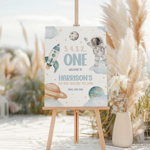 May include: A blue and white sign with a rocket, astronaut, planets, and stars. The sign says "5, 4, 3, 2... ONE Welcome to Harrison's 1st Trip Around the Sun! April 10th 2026".
