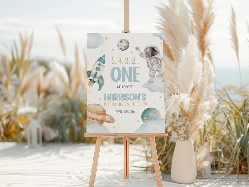 May include: A blue and white sign with a rocket, astronaut, planets, and stars. The sign says "5, 4, 3, 2... ONE Welcome to Harrison's 1st Trip Around the Sun! April 10th 2026".
